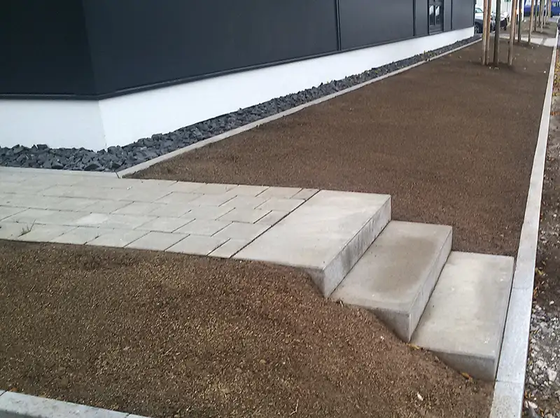 Frisch angelegte Außenanlage eines modernen Gewerbegebäudes von PUR Projektbau mit gepflastertem Gehweg, Betonstufen und vorbereitetem Erdboden für die Bepflanzung.