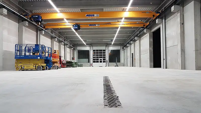 Innenansicht einer großen, hellen Industriehalle mit Betonboden und Deckenkran.