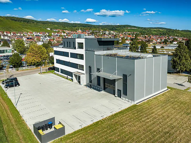 Luftaufnahme eines Gewerbebaus mit großem Vorplatz und umliegender Landschaft im Hintergrund.