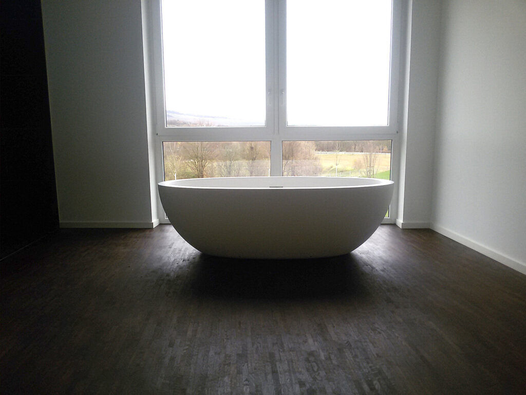 Moderne, freistehende Badewanne auf dunklem Holzboden in einem lichtdurchfluteten Gebäude.