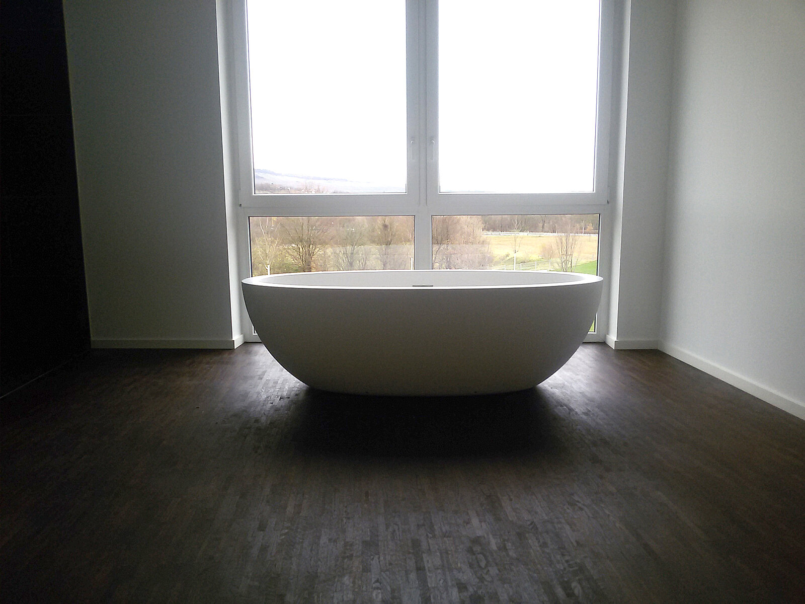 Moderne, freistehende Badewanne auf dunklem Holzboden in einem lichtdurchfluteten Gebäude.